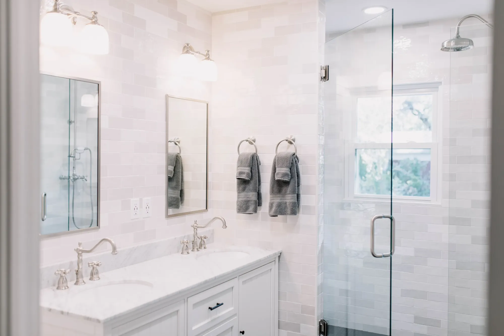 A white double vanity with a veined white stone top, wall sconces, and a clear glass shower under a window in the Arlington Heights primary bath