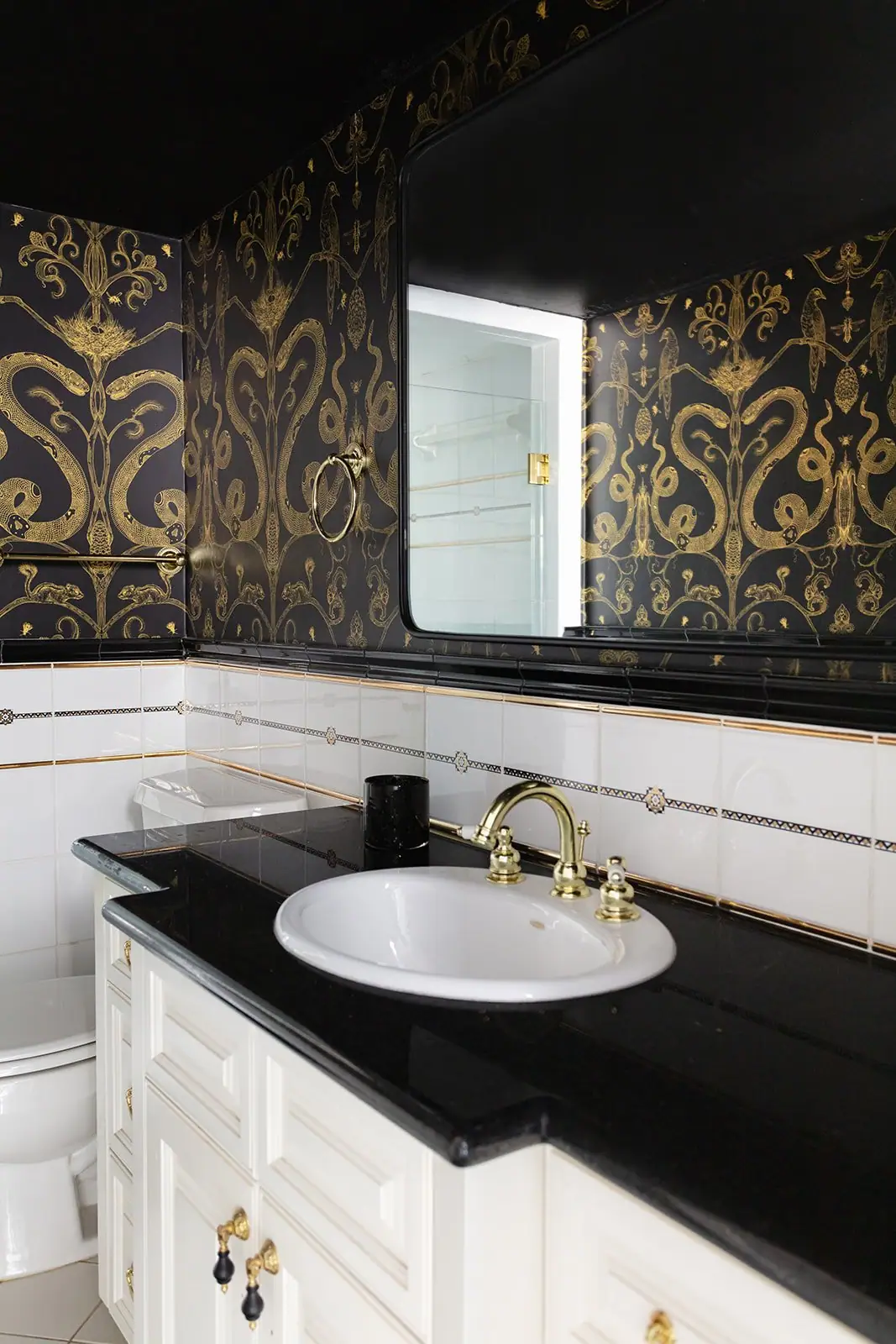 A brass faucet on a black counter with white tile and black-and-gold patterned wallpaper in a Mistletoe Heights bath