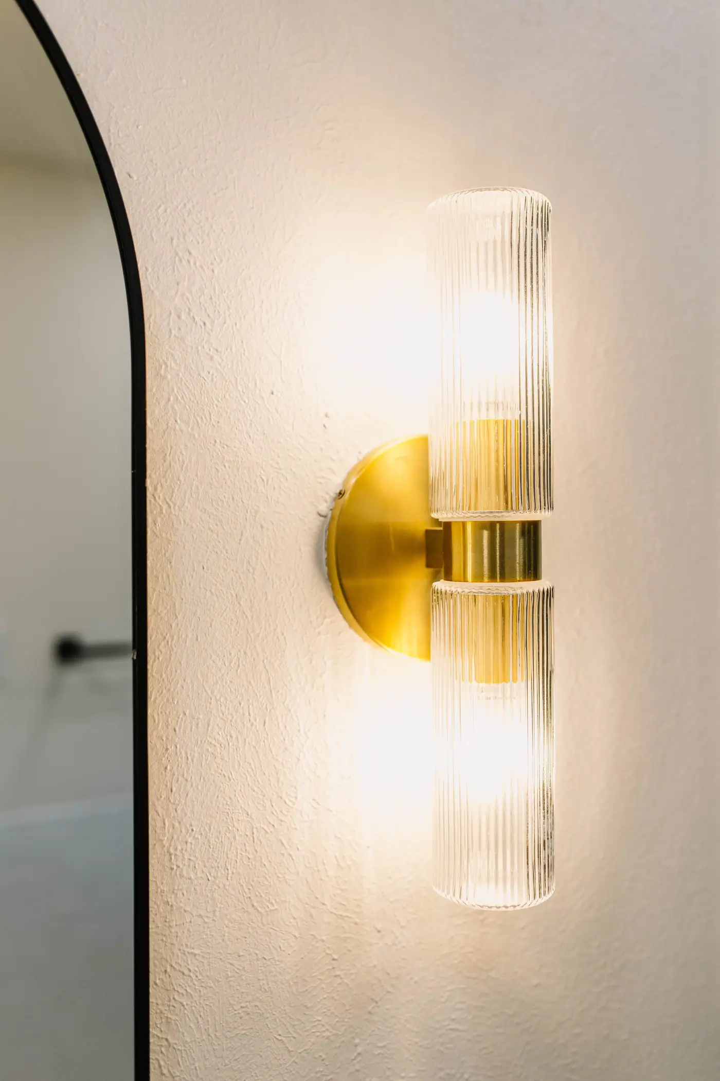 A brass sconce with two ribbed glass shades beside the curved black edge of the arched mirror in the Overton Park primary bath