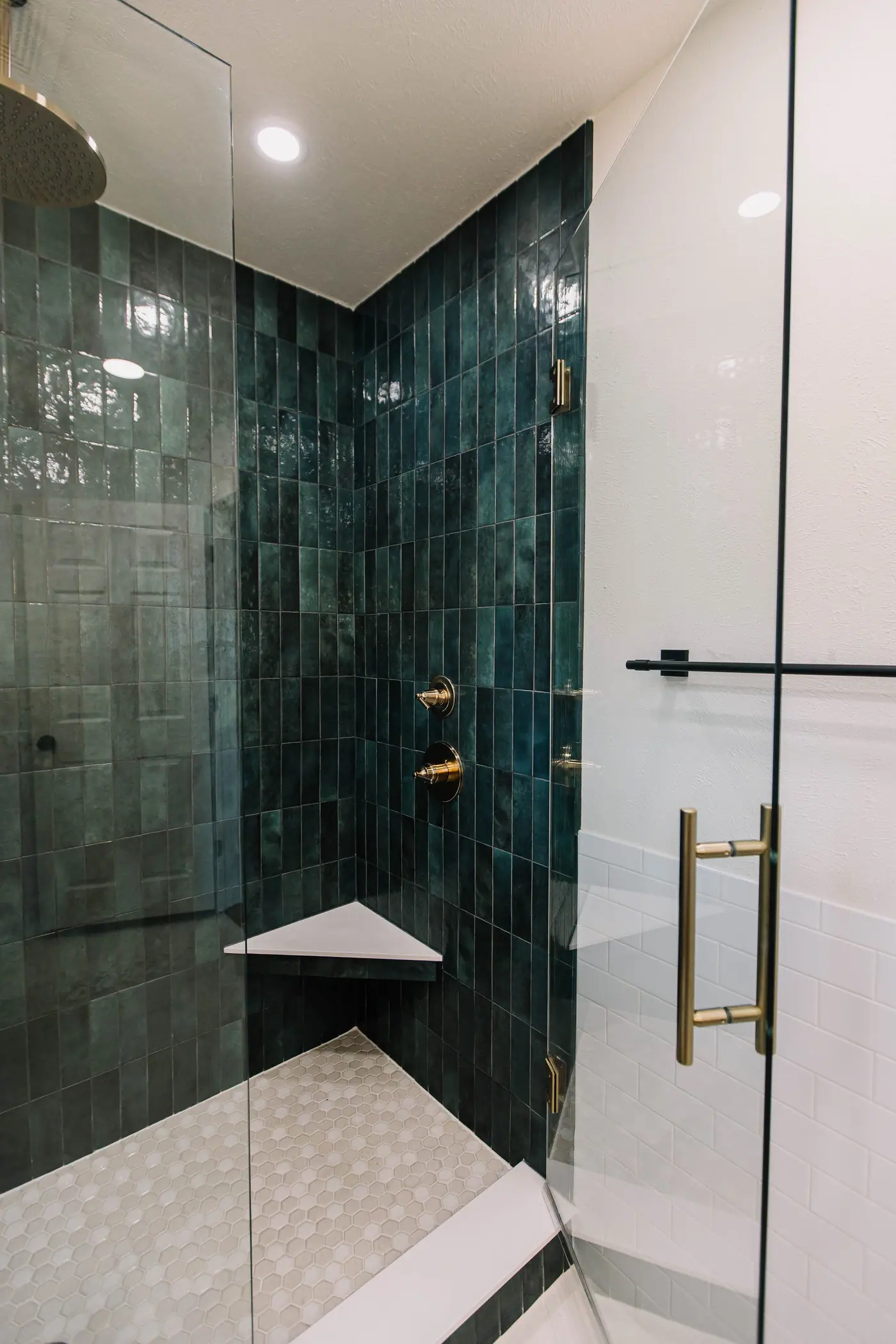 A corner bench and white hexagon mosaic floor in a deep green tile shower with champagne bronze valves and pull in the Overton Park primary bath