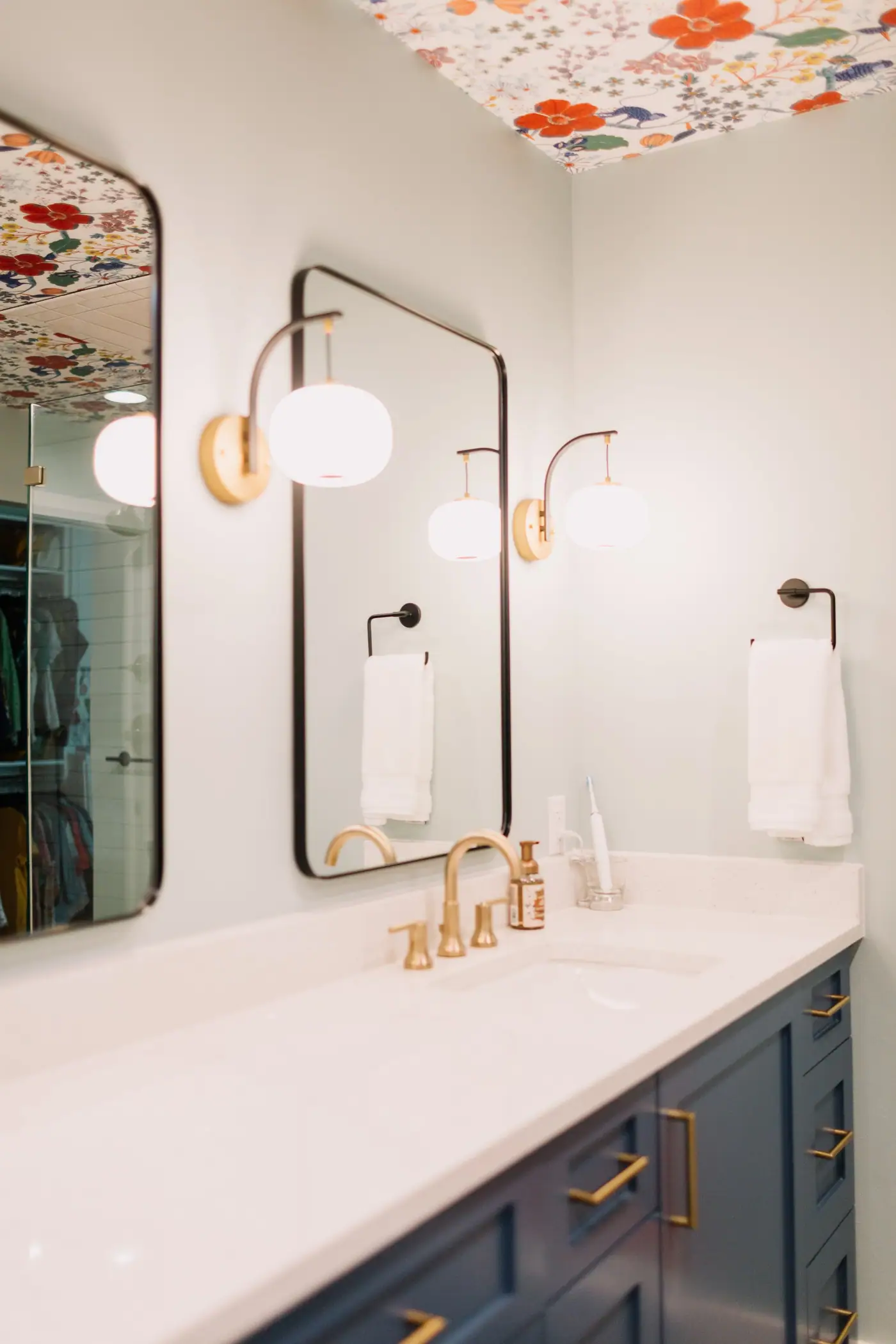 A navy double vanity with brass pulls and brass faucets under black-framed mirrors and brass sconces, beneath a floral papered ceiling, in an Overton Park primary bath