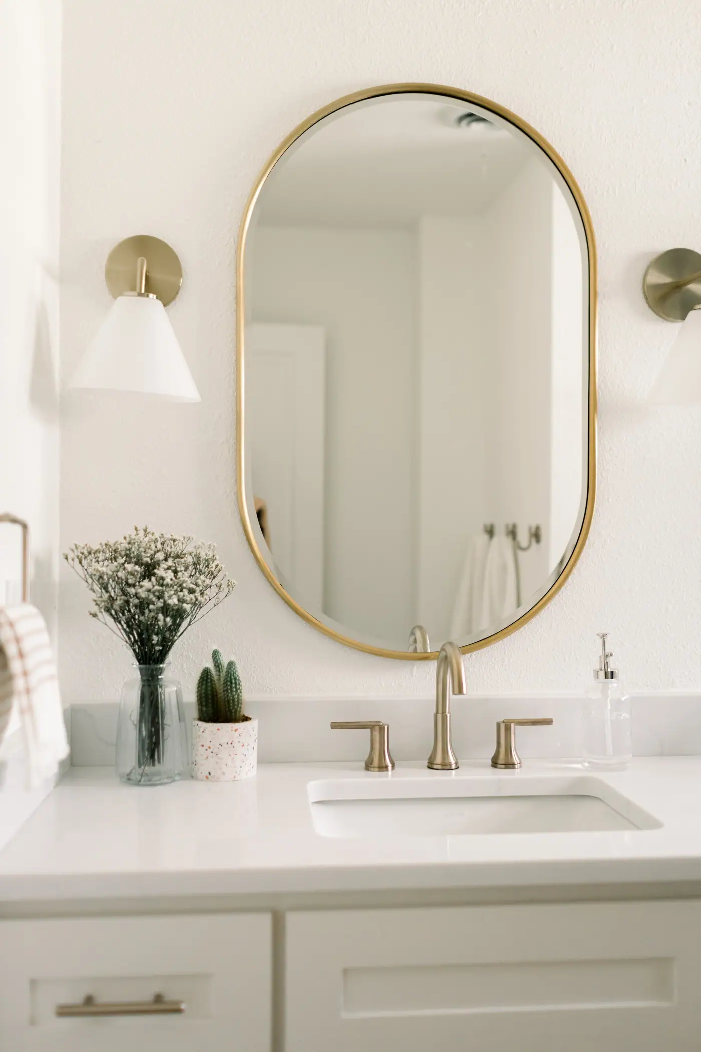 An oval brass-framed mirror over a gold widespread faucet, a white cone sconce, and a vase of baby's breath on the counter in a Ridglea Hills Addition bath
