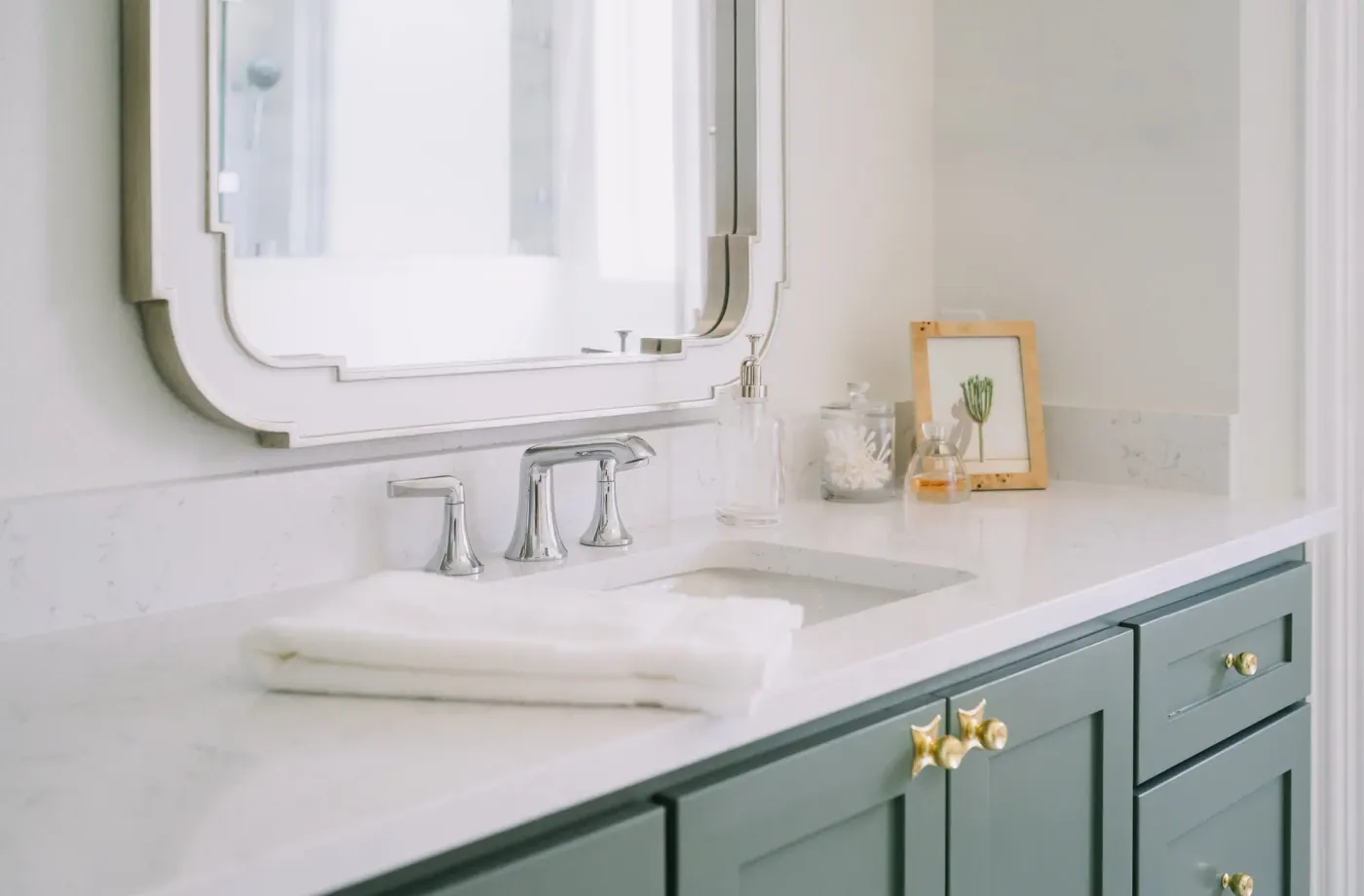 A sage green vanity and tall linen cabinet with brass knobs, under a brass sconce, in the Riverwood bath refresh