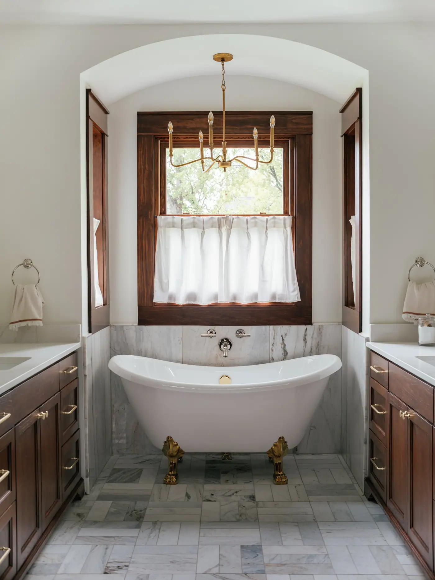 A clawfoot tub on brass feet between twin stained wood vanities under an arched ceiling and a brass chandelier in a Ryan Place addition bath