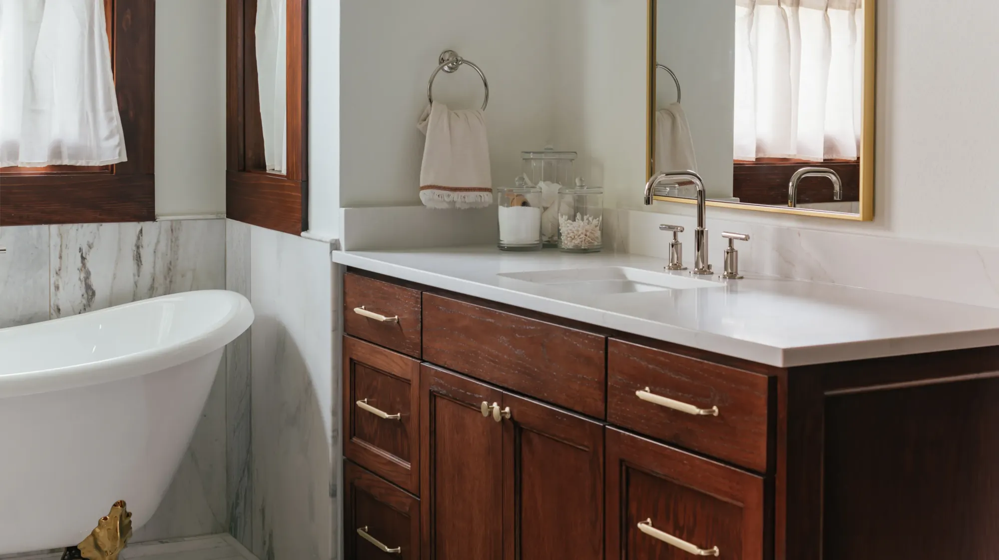 A stained wood vanity with brass pulls under a brass-framed mirror and a milk-glass sconce, beside a clawfoot tub on brass feet and marble wainscot, in a Ryan Place primary bath