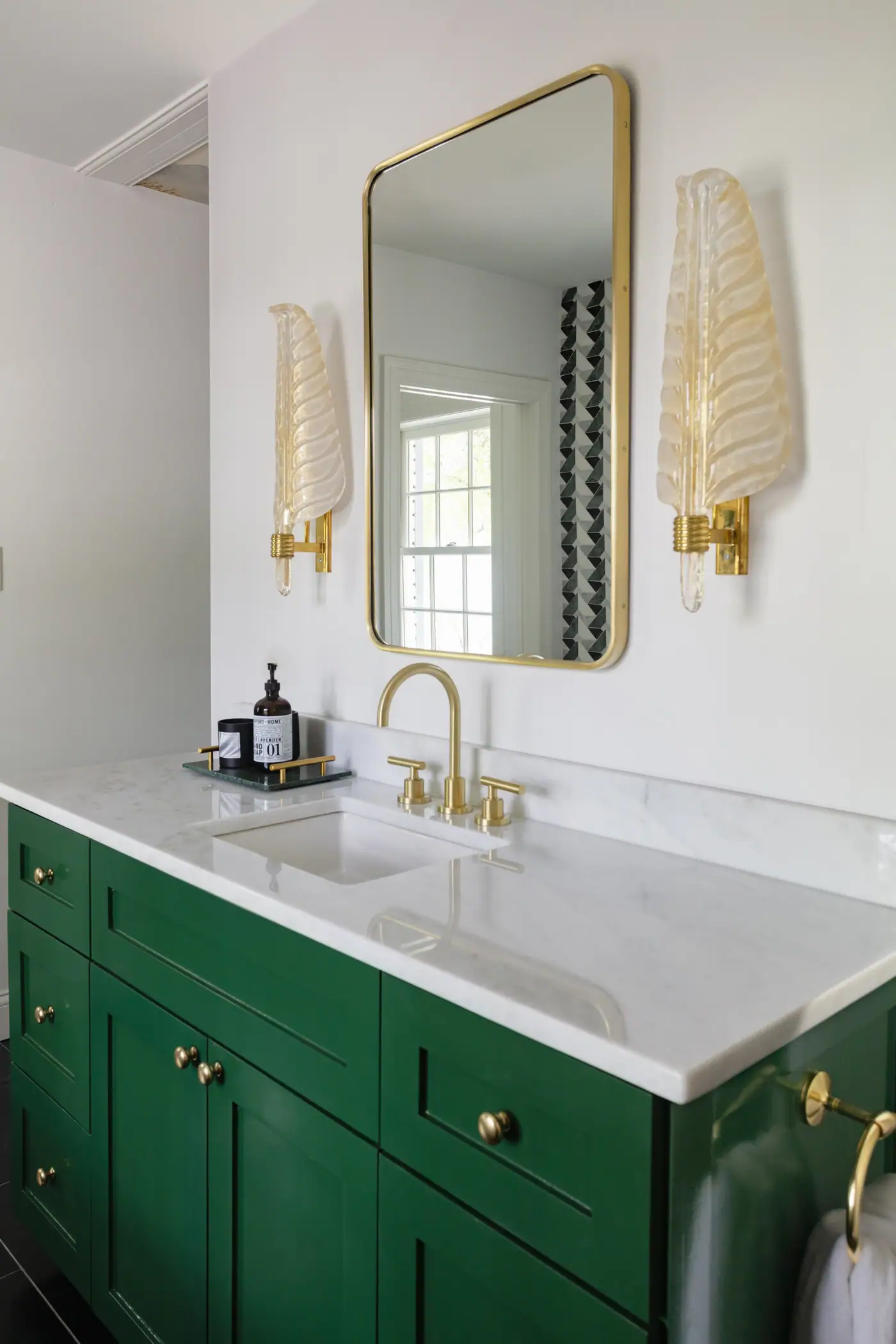 Glass leaf sconces flank a brass-framed mirror above a green vanity with a veined white stone top in the Tanglewood primary suite