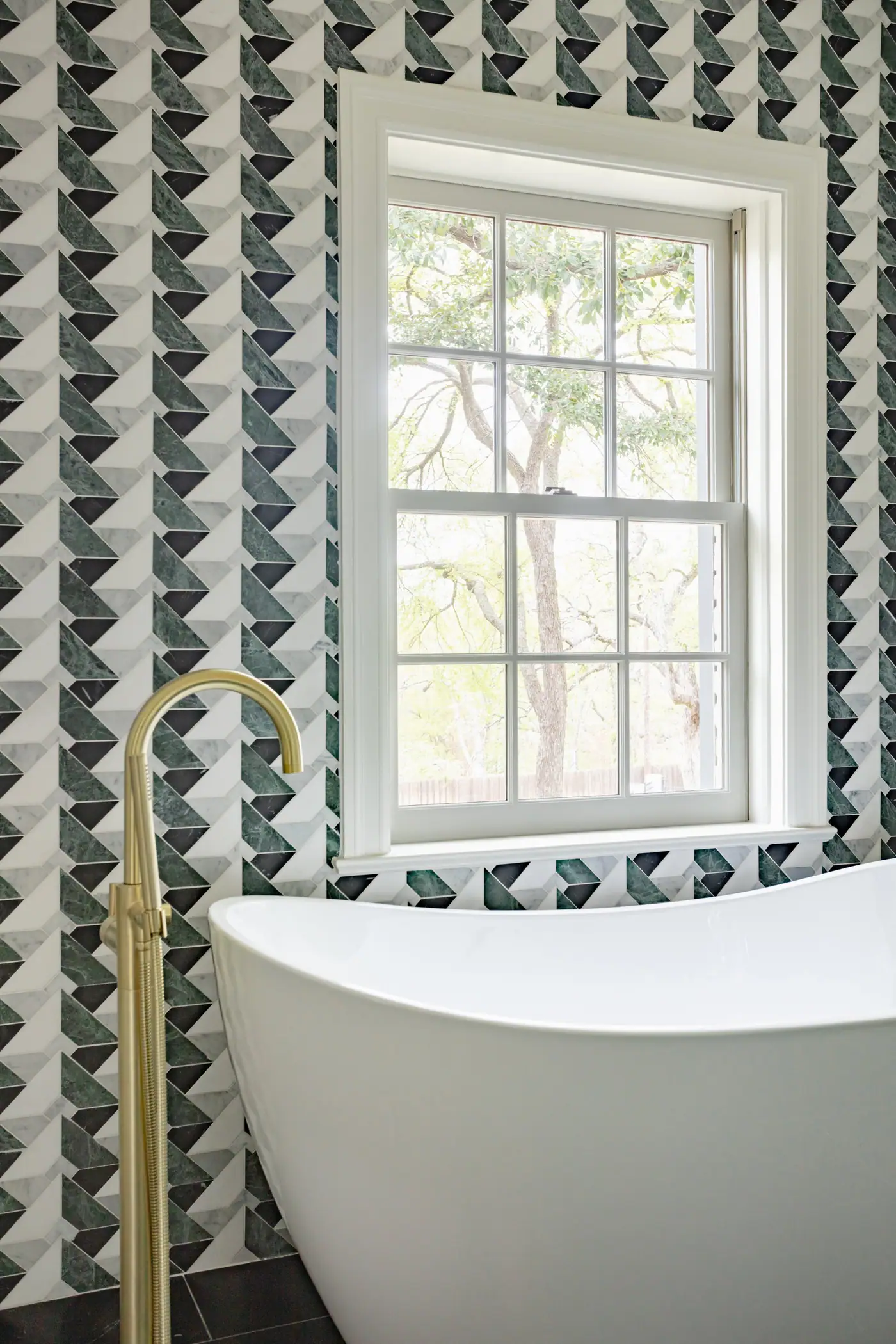 A brass floor-mounted tub filler and freestanding tub under a window framed in green and black stone mosaic in the Tanglewood primary suite