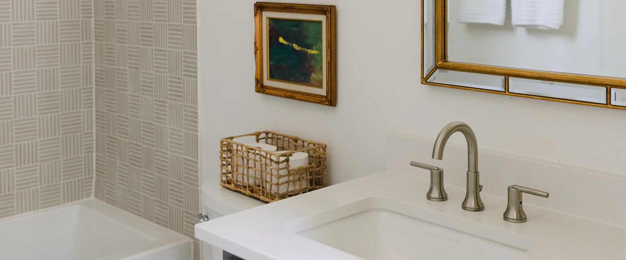 A blue-grey vanity with a gold bamboo mirror and globe sconces, beside a basketweave tiled tub