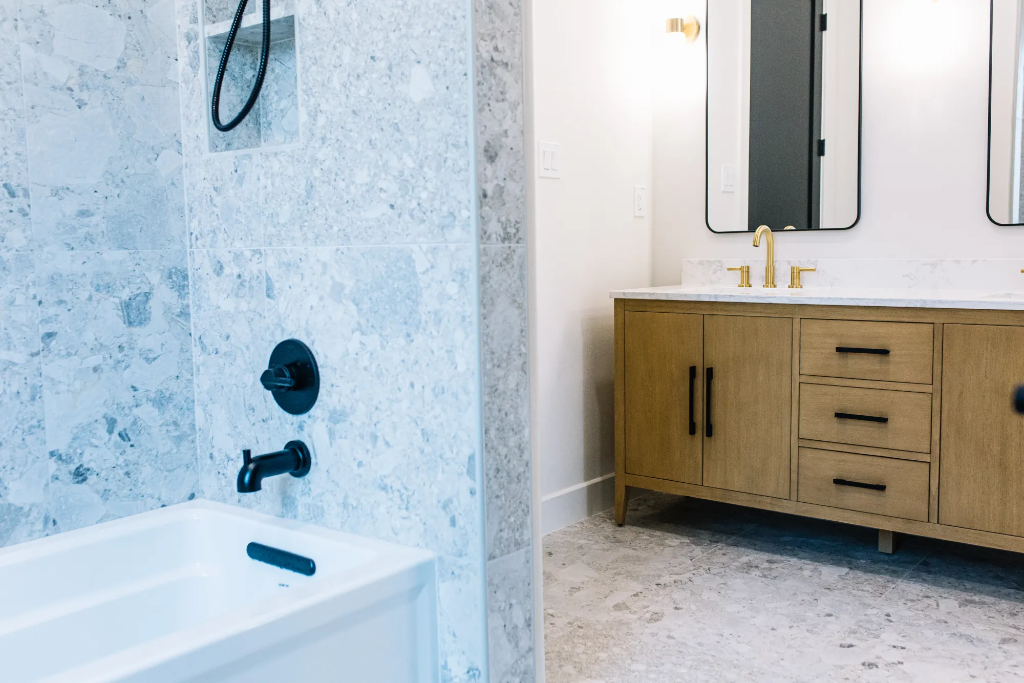A blue-grey terrazzo tub surround with matte black fittings, across from an oak double vanity