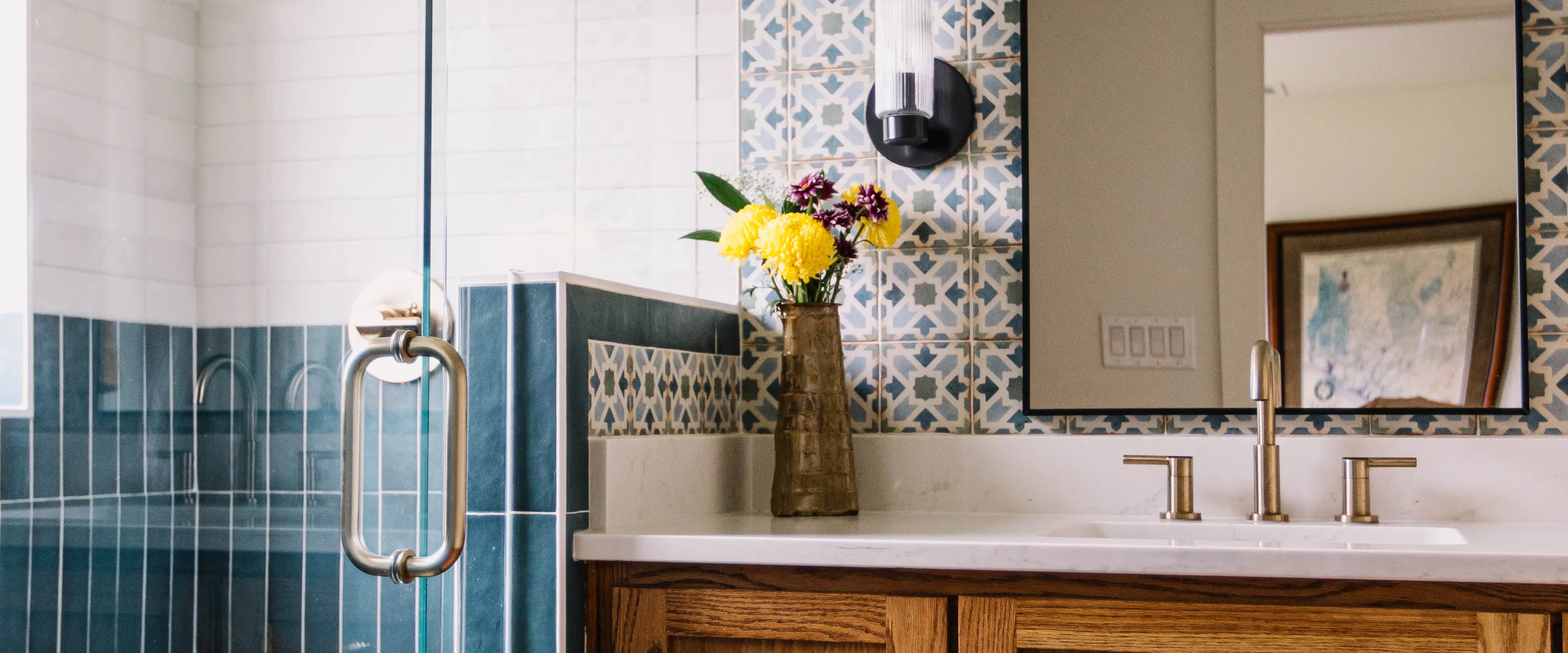 A blue vertical-stack tiled shower behind frameless glass beside blue and white patterned wall tile, a black sconce and a black-framed mirror, above a white quartz counter with a champagne bronze faucet