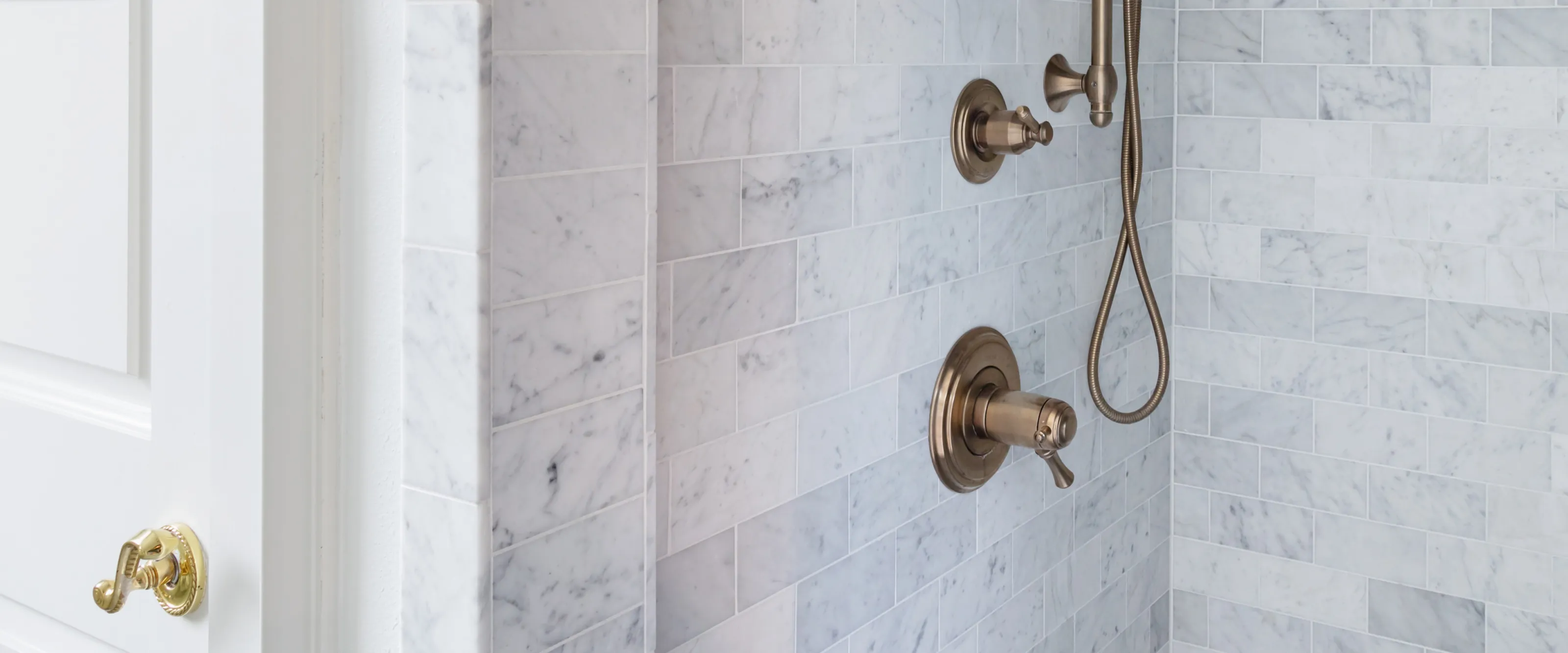 A Carrara marble subway tiled shower with an antique brass valve and a hand shower on its hose, beside a white panelled door