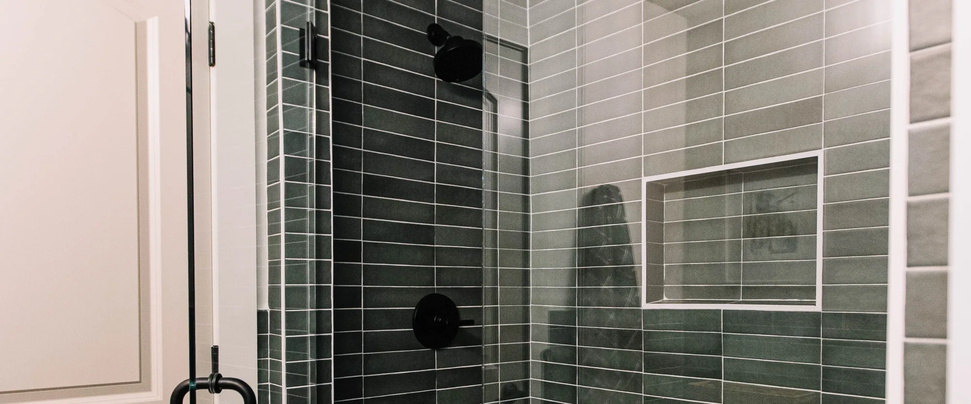 A green tiled shower with matte black fixtures and a penny-round floor, behind glass on patterned tile