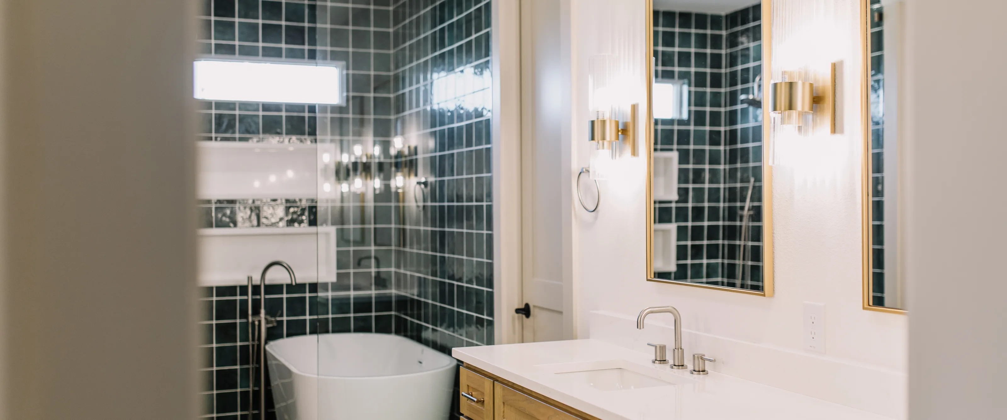 A freestanding tub in a green square tiled wet room behind glass, beside an oak vanity with a white quartz top under brass-framed mirrors and fluted brass sconces