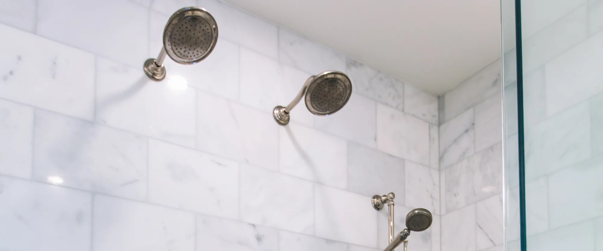 A marble tiled shower with twin nickel heads, a hand shower on a slide bar, a marble bench, and a blue-grey fish-scale mosaic floor