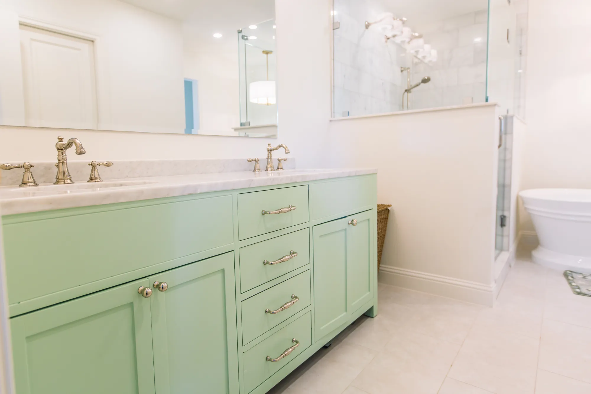 A mint green double vanity with a marble top and nickel bridge faucets, beside a marble tile shower