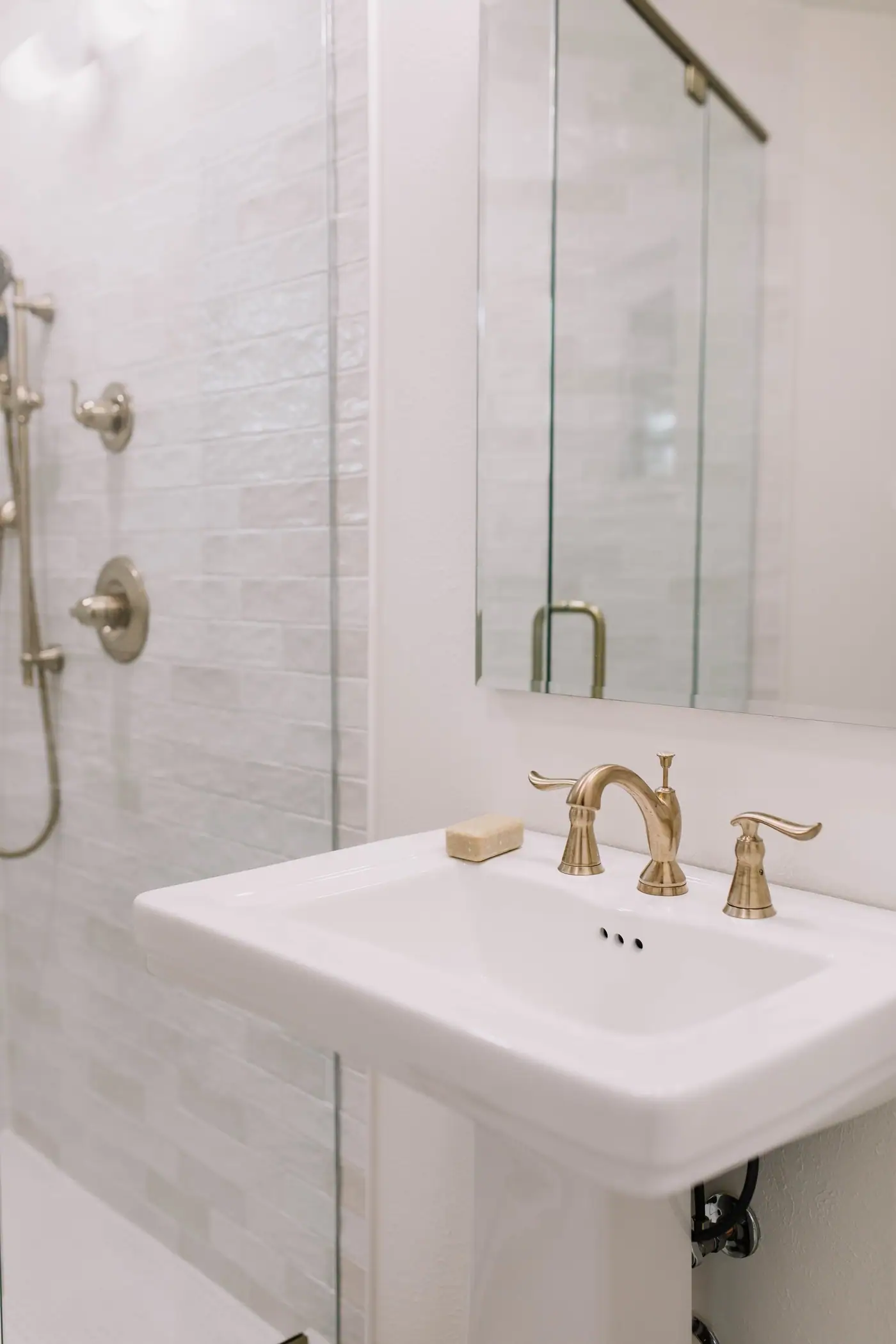 A white pedestal sink with a brass widespread faucet beside a white tile shower with nickel-toned fixtures in a Mistletoe Heights bath
