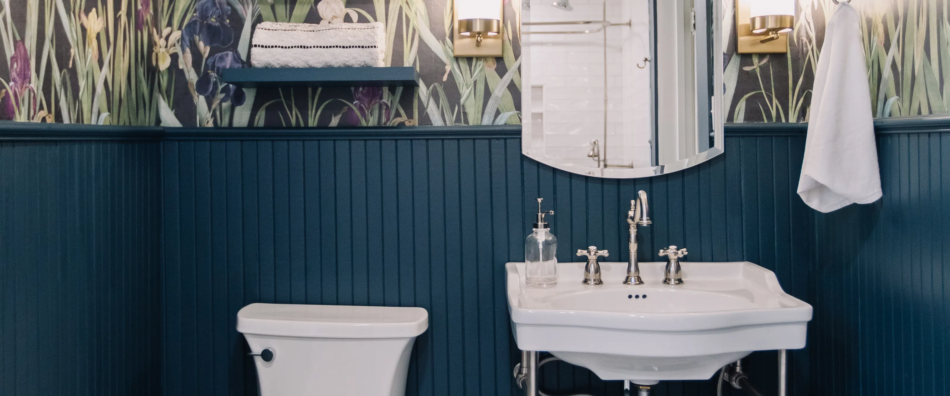 A white console sink on metal legs with a cross-handle bridge faucet, under an arched mirror between two brass sconces, against navy beadboard below iris-print wallpaper