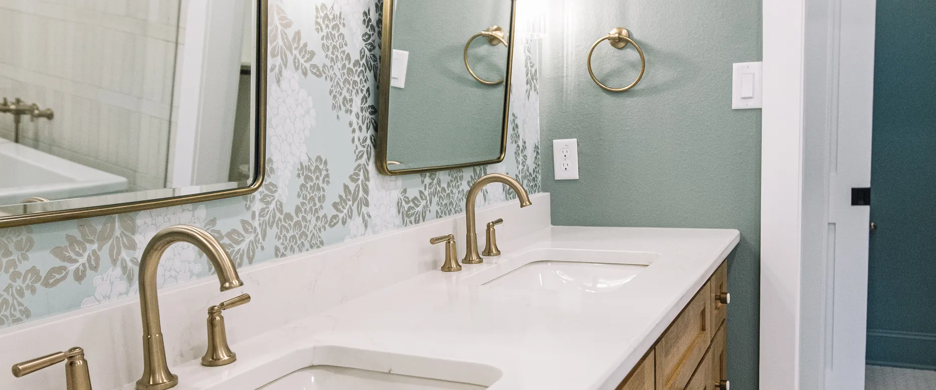 A white oak double vanity with brass faucets and a white quartz top under twin brass-framed mirrors, against blue-green floral wallpaper