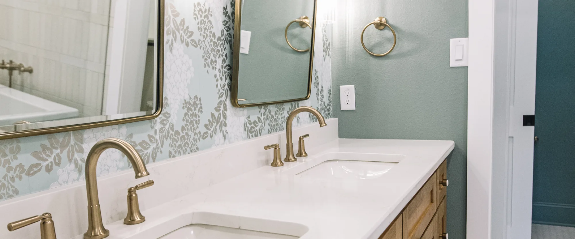 A white oak double vanity with brass faucets under twin arched brass mirrors and crystal sconces, against blue-green floral wallpaper