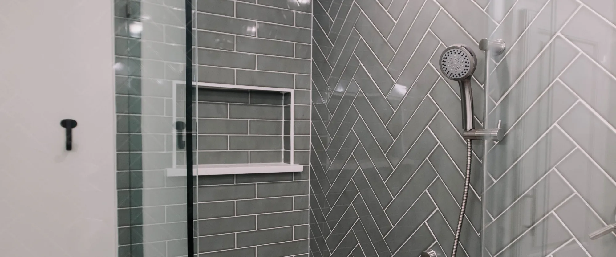 A slate green herringbone tiled shower with a marble bench and a nickel hand shower, behind frameless glass