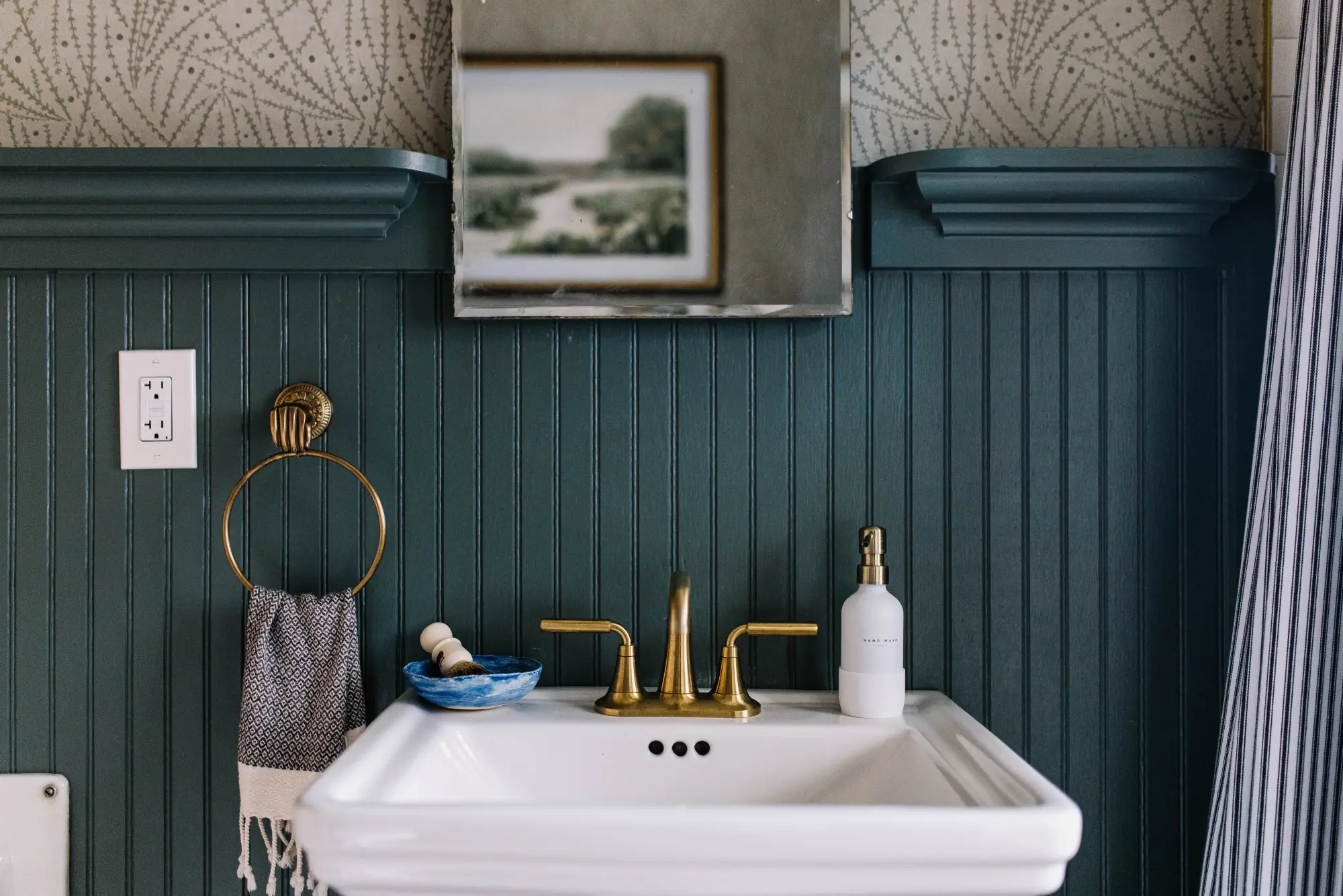 A white pedestal sink with a brass faucet against deep teal beadboard, below botanical wallpaper and an antique mirror