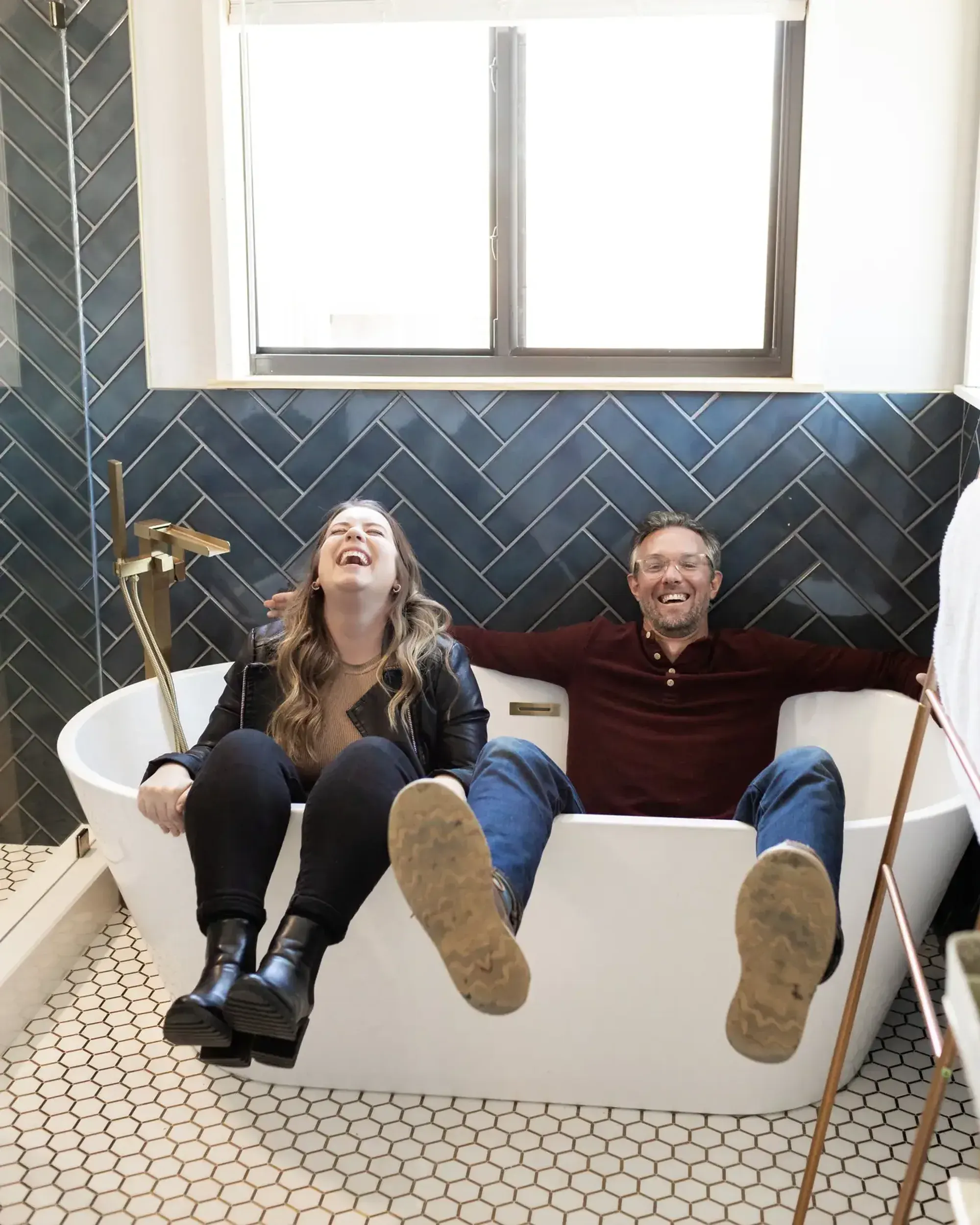 Two 6th Avenue Homes team members sitting in a newly installed freestanding tub, laughing, against a navy herringbone tile wall